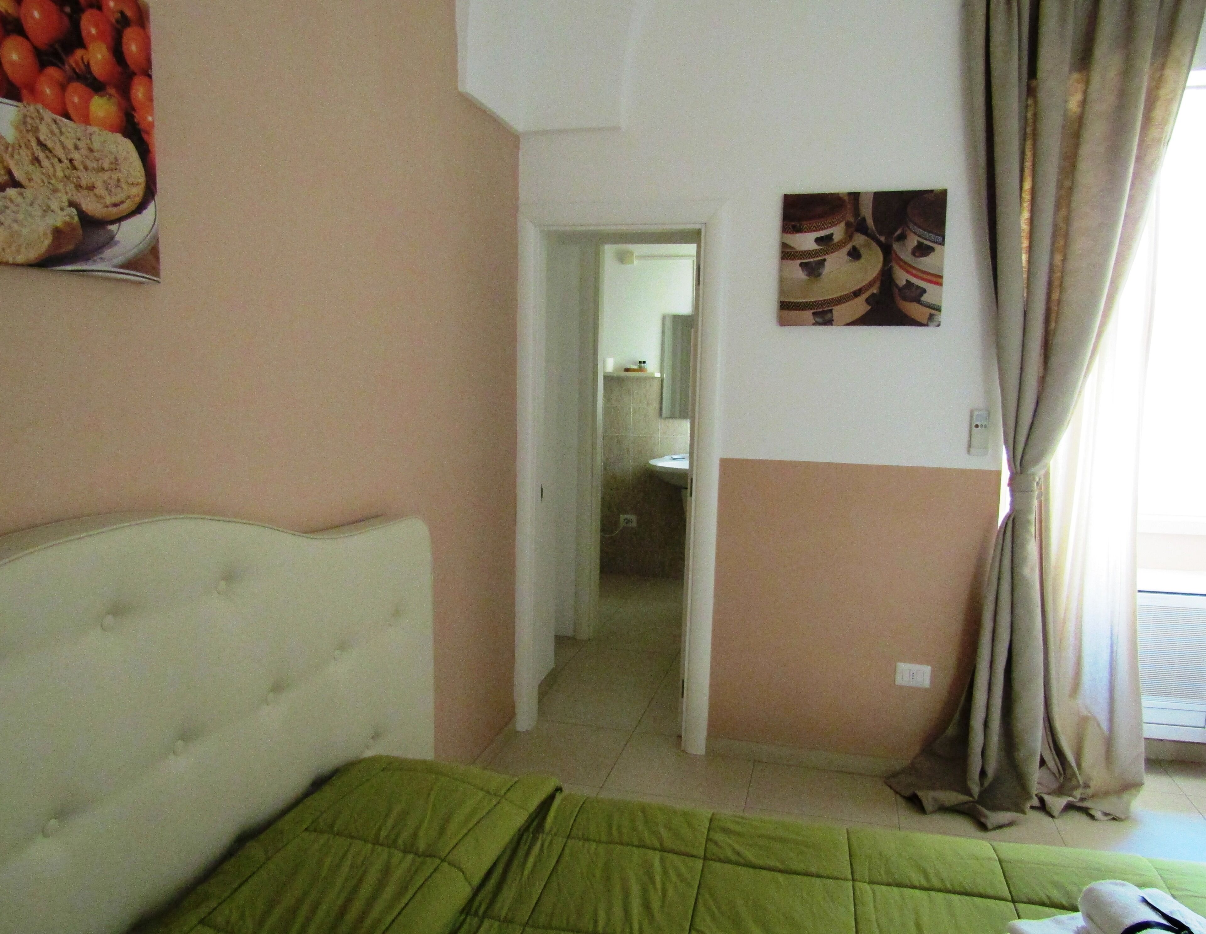 A hotel room with a bed, a green bedspread, a wall with a fruit picture, a door leading to a bathroom, and a window with curtains.