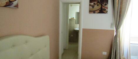 A hotel room with a bed, a green bedspread, a wall with a fruit picture, a door leading to a bathroom, and a window with curtains.