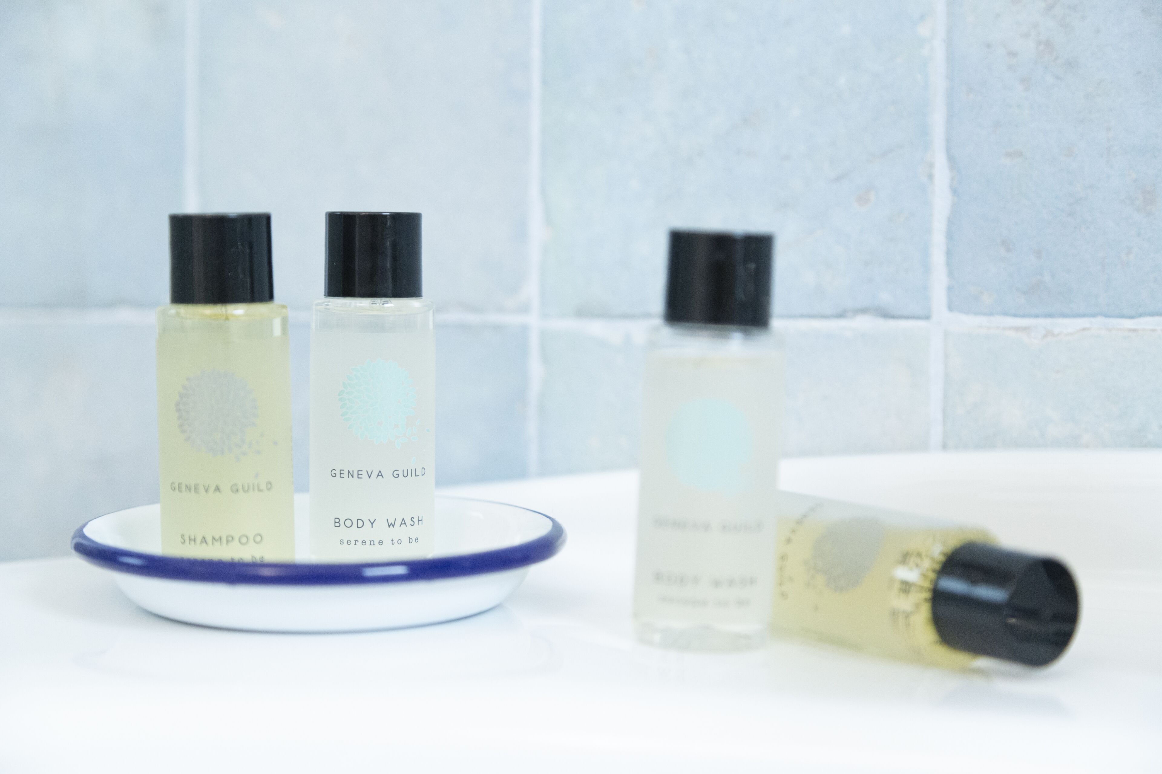 Three bottles of Geneva Guild body wash and shampoo on a white surface with a blue rimmed dish.