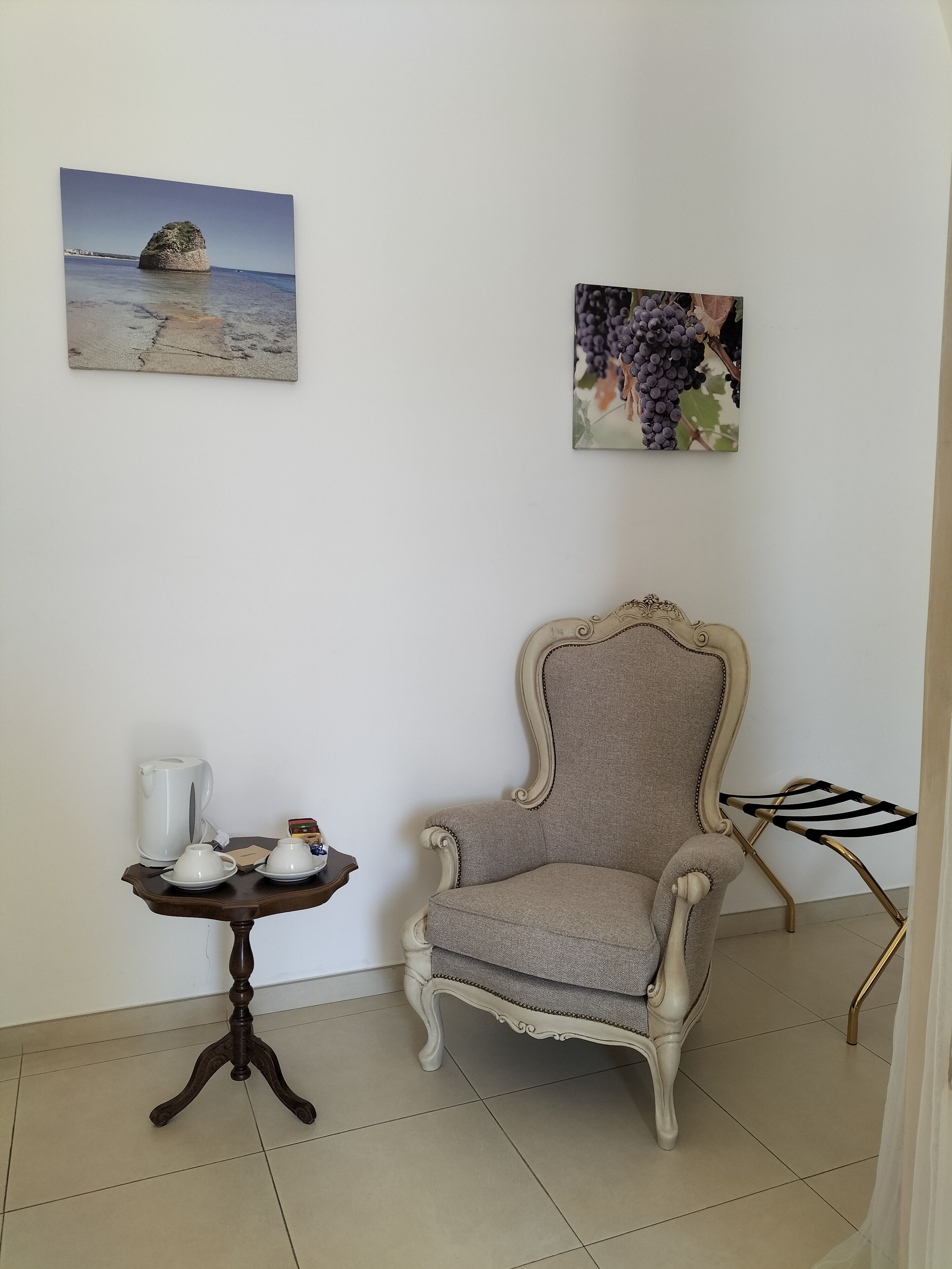 Une pièce comprenant un fauteuil classique, une petite table avec une bouilloire et des tasses, ainsi que deux œuvres d’art accrochées au mur, représentant une plage et des raisins.