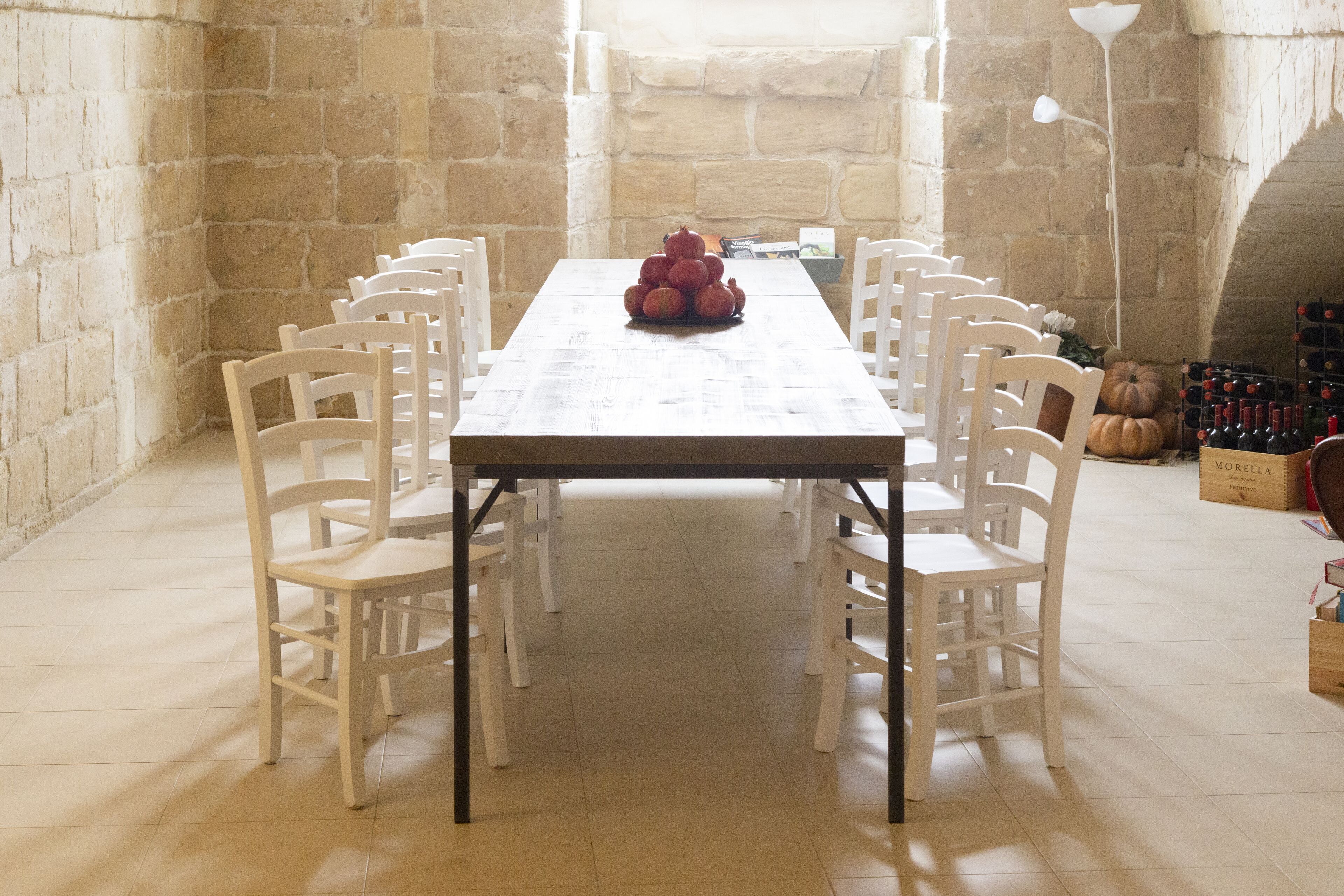 Une longue table à manger entourée de chaises blanches, une composition de pommes au centre et un meuble à vin en arrière-plan.
