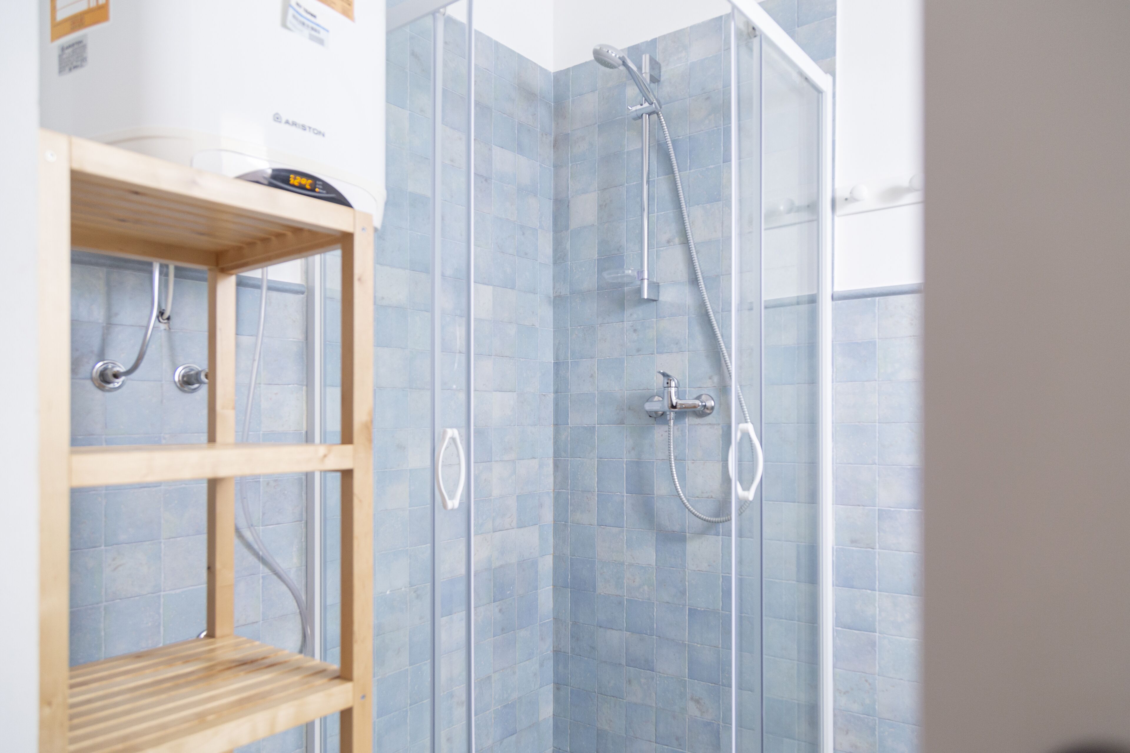 Une douche équipée d’un pommeau de douche à main et d’un robinet mural, d’une étagère en bois et d’un mur recouvert de carreaux.