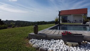A pool with a wooden deck, a house with a red roof, and a grassy landscape with hills in the background. - Cottage between sea and mountains with private pool (and heated) - View Pyrenees (Laà-Mondrans)