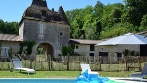 Une piscine entourée de chaises longues et de jeux gonflables, le tout délimité par une clôture en bois et bordé d’un bâtiment en pierre doté d’un toit en tuiles.