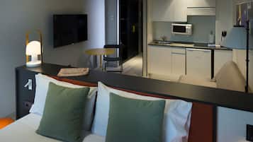 Habitació d'hotel amb un llit, almoades verdes i blanques, una taula de nit amb una làmpada, una televisió, una taula rodona petita i una cuineta amb microones.