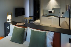 A hotel room with a bed, green and white pillows, a bedside table with a lamp, a television, a small round table, and a kitchenette with a microwave. - The Oliver Apartamentos Pozuelo (Pozuelo de Alarcon)
