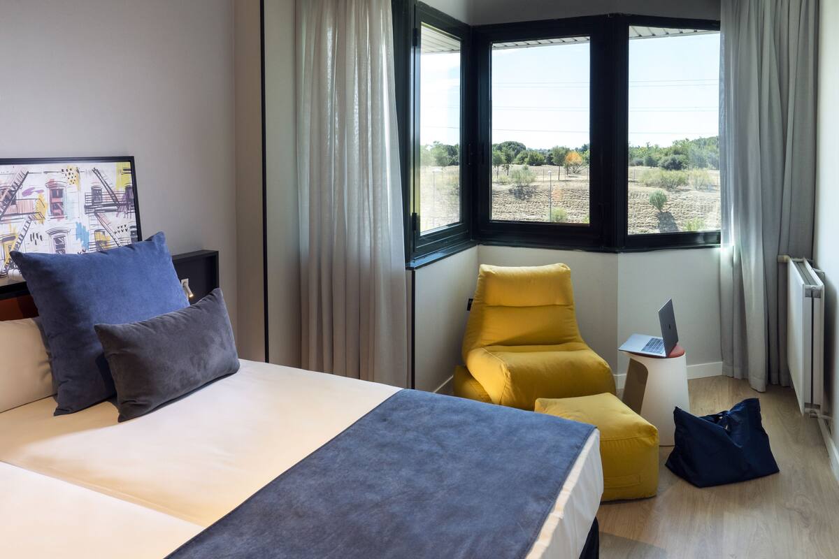 Ein Hotelzimmer mit Bett, einem gelben Sessel, einem Laptop auf einem kleinen Tisch und Blick ins Freie.