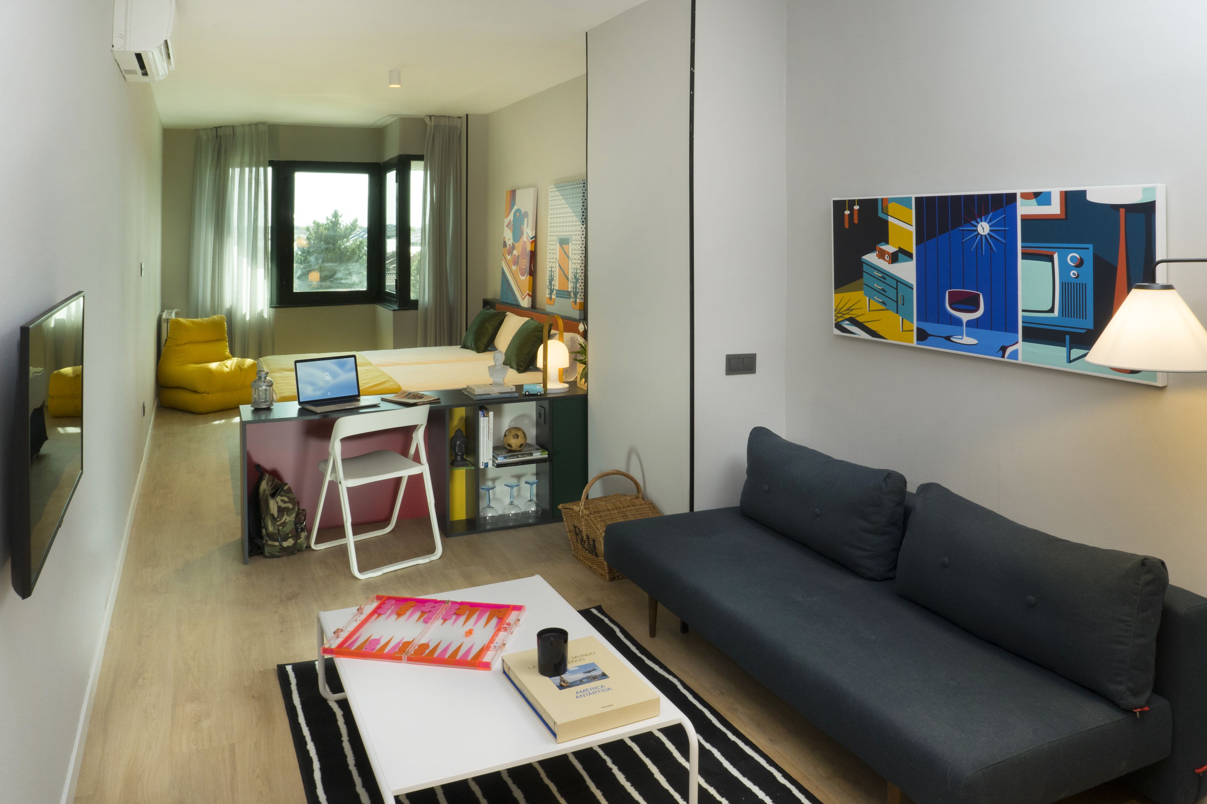 Una habitación de hotel moderna con sofá, escritorio con computadora portátil, cama con vista y un cuadro en la pared.