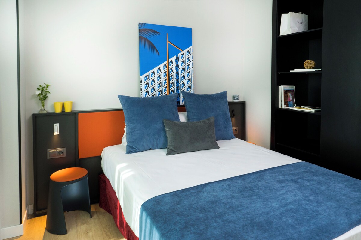 Una camera d'albergo moderna con un letto, un comodino, un tavolino con un vaso e due tazze, una libreria e un quadro appeso alla parete.