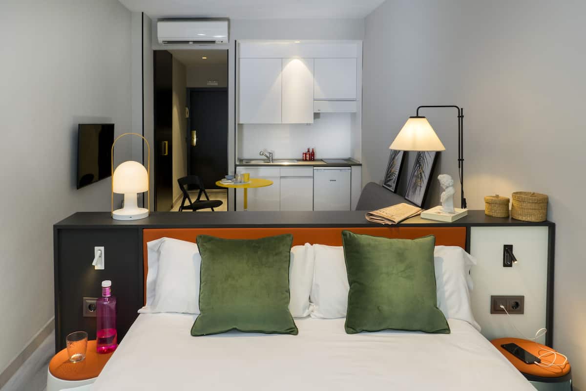 Ein Hotelzimmer mit Bett, Nachttisch, Lampe und Blick auf den Küchenbereich.