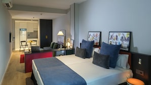 A modern hotel room with a large bed, a desk with a laptop, a chair, and a small table. There are two framed pictures on the wall and a lamp on the desk.