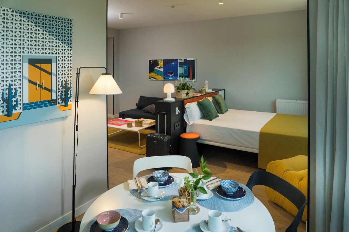 Habitación de hotel con cama, escritorio, silla, mesa dispuesta para el desayuno y una pared con obras de arte.