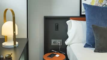 A hotel room with a bed, a nightstand with a lamp, a small table with a phone and glasses, and a backpack on the floor.