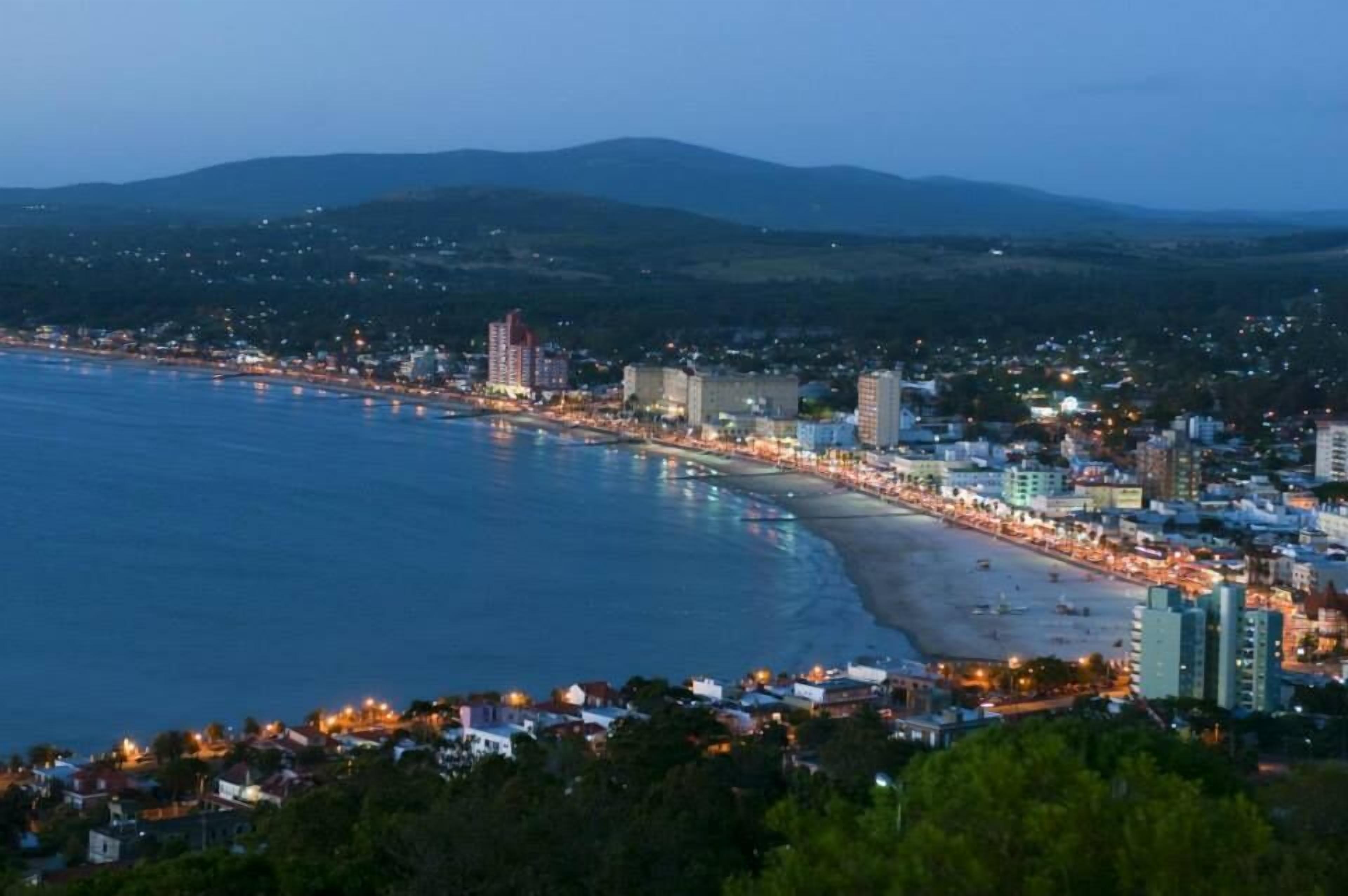 Un paisaje costero con playa, edificios y una cadena montañosa al fondo.