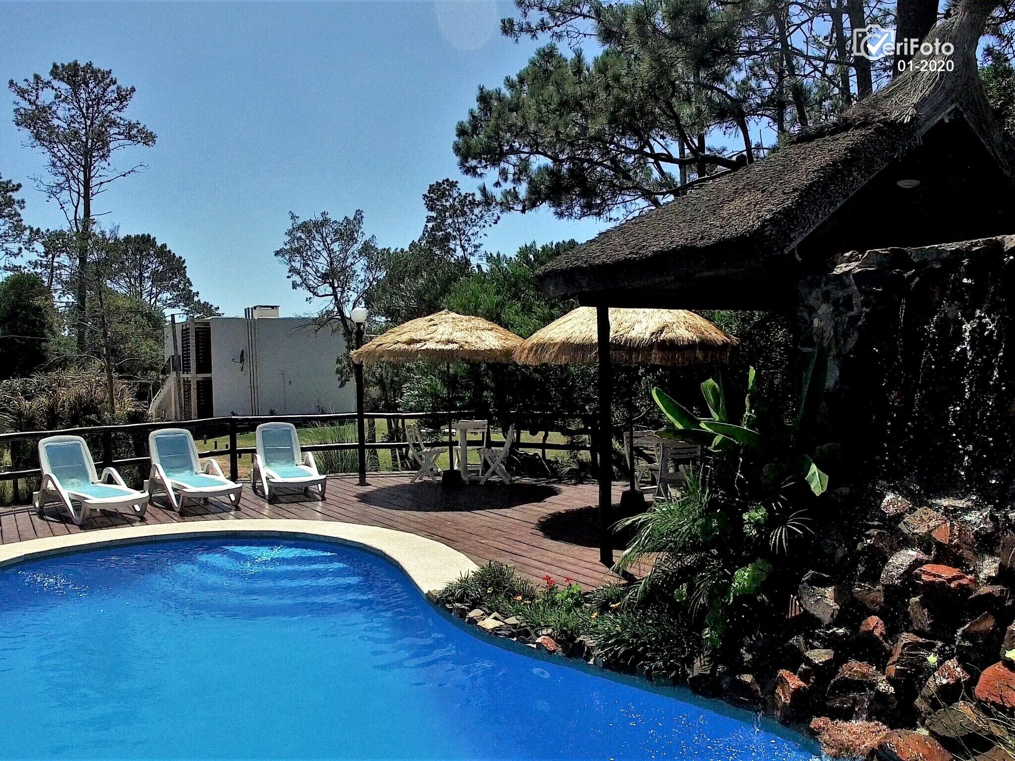 Piscina con sillones de descanso y sombrillas de paja, rodeada de vegetación y un muro de piedra.