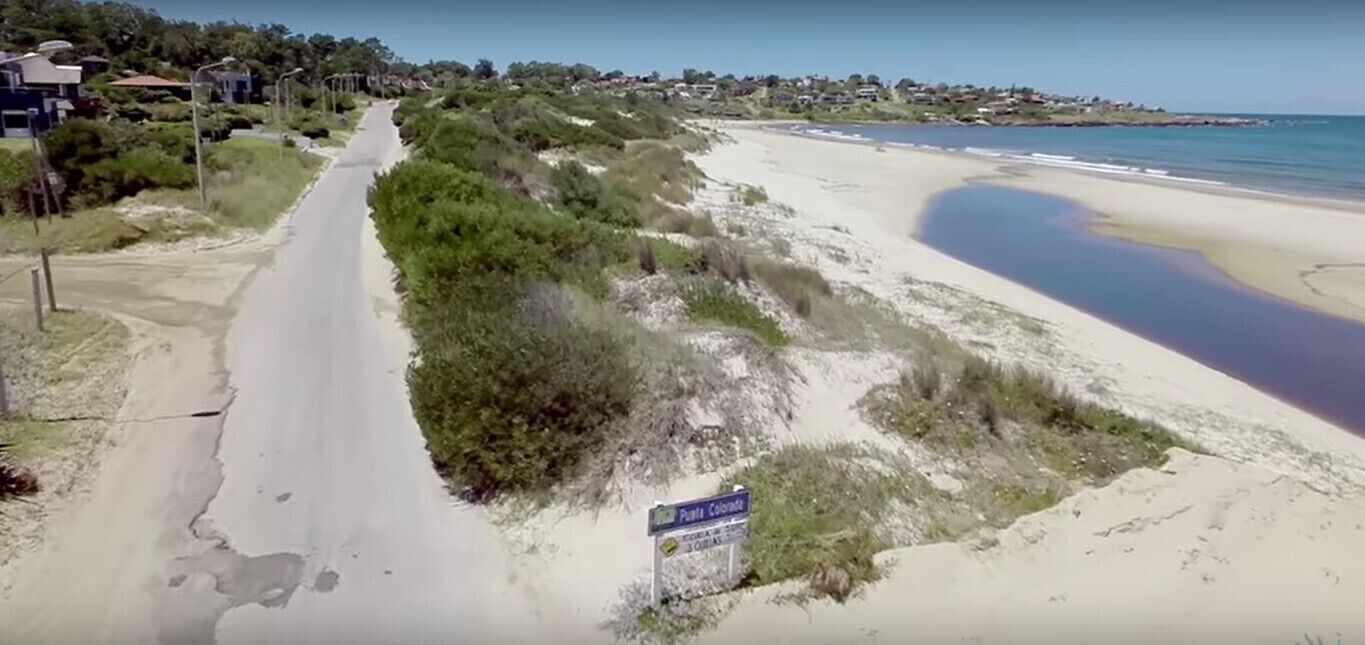 Una carretera costera con una playa a la derecha y casas al fondo.