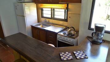 Une cuisine compacte avec des meubles en bois, un four à micro-ondes encastré, une cuisinière à gaz, une machine à café et une fenêtre avec des stores.