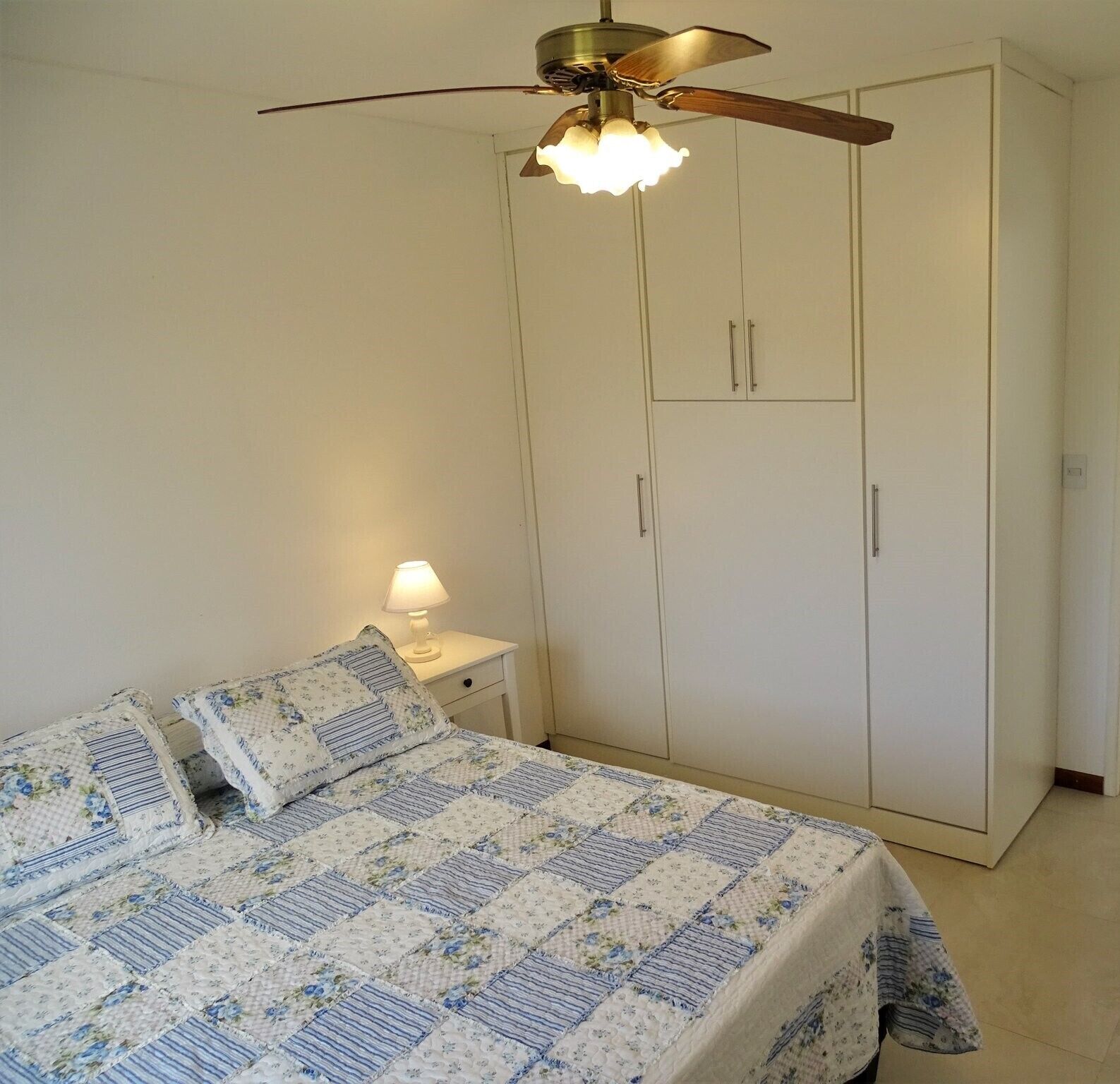 A bedroom with a bed, nightstand, lamp, wardrobe, and ceiling fan.