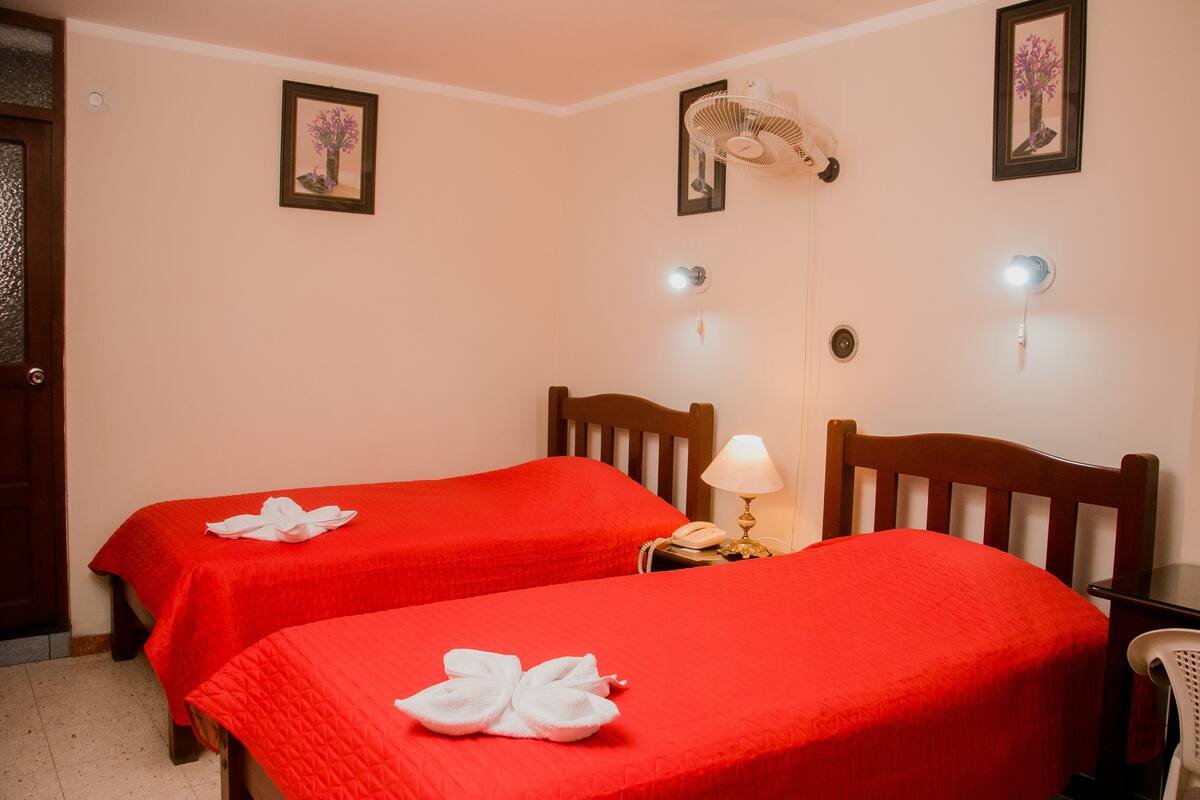 A hotel room with two beds, each with a red bedspread and a white towel. There are wall-mounted fans and picture frames on the walls.