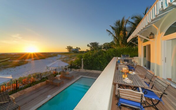 A poolside area with a view of the sunset over the water, a dining table with chairs, and a covered seating area.