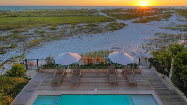 A poolside area with lounge chairs and umbrellas, overlooking a beach and ocean at sunset. - Villa Marisol: Beachfront, Mediterranean style, w/terrace, pool, hot tub! (Anna Maria)