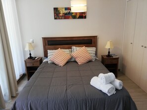 A neatly made bed with a dark grey duvet, two bedside tables with lamps, a painting on the wall, and a wardrobe. - Apartment near the center. New, very welcoming. (Pucon)