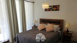 Un lit bien fait, recouvert d’une couverture marron, avec deux oreillers et des serviettes enroulées au pied du lit. On y trouve deux tables de chevet avec des lampes, et un tableau accroché au mur au-dessus du lit.