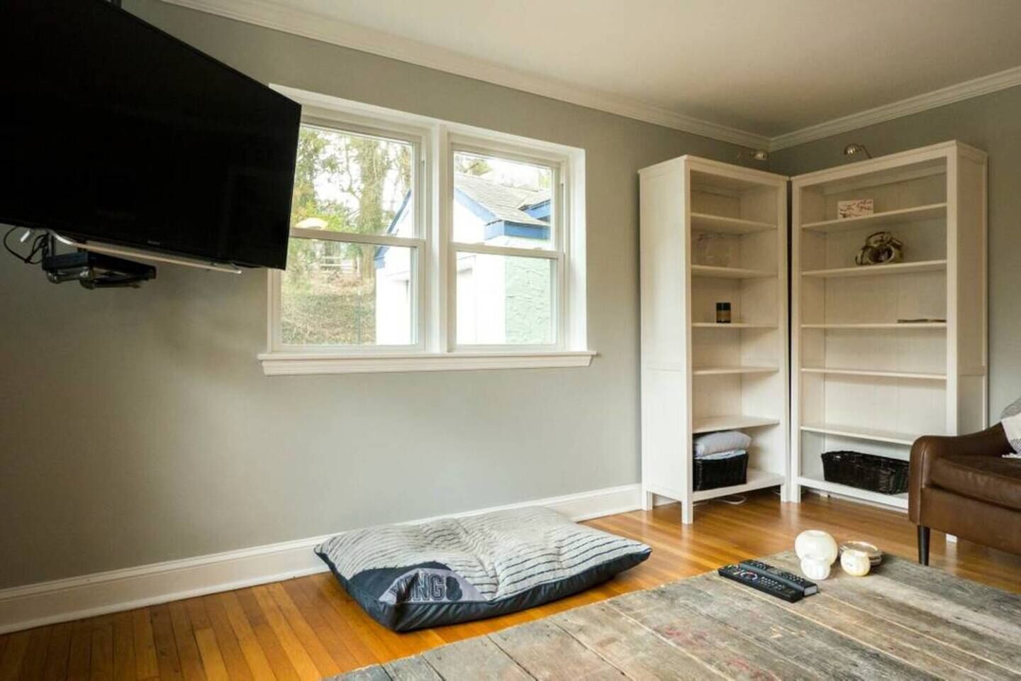 Una stanza con una grande finestra, una TV fissata al muro, un pavimento in legno, una poltrona in pelle marrone e un mobile contenitore bianco.
