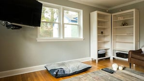 Ein Zimmer mit einem großen Fenster, einem an der Wand befestigten Fernseher, Holzboden, einem braunen Ledersessel und einer weißen Regaleinheit.