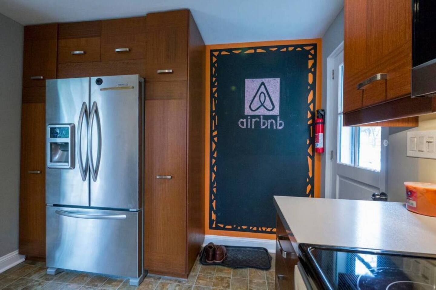 A compact kitchen with a stainless steel refrigerator, wooden cabinets, and a door with an Airbnb logo.