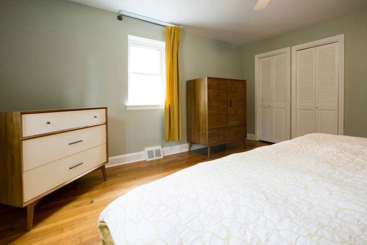 A bedroom with a bed, a dresser, a wardrobe, a window with curtains, and a ceiling fan.