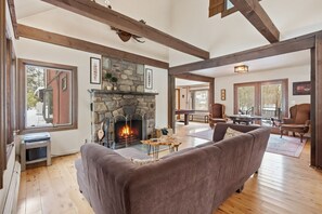 A cozy living room with a stone fireplace, wooden beams, and a large window overlooking a snowy landscape. - Private 10-Acre Creekside Cabin—80-Ft Deck, Fire Pit, Wildlife & Cozy Spaces! (Canadensis)