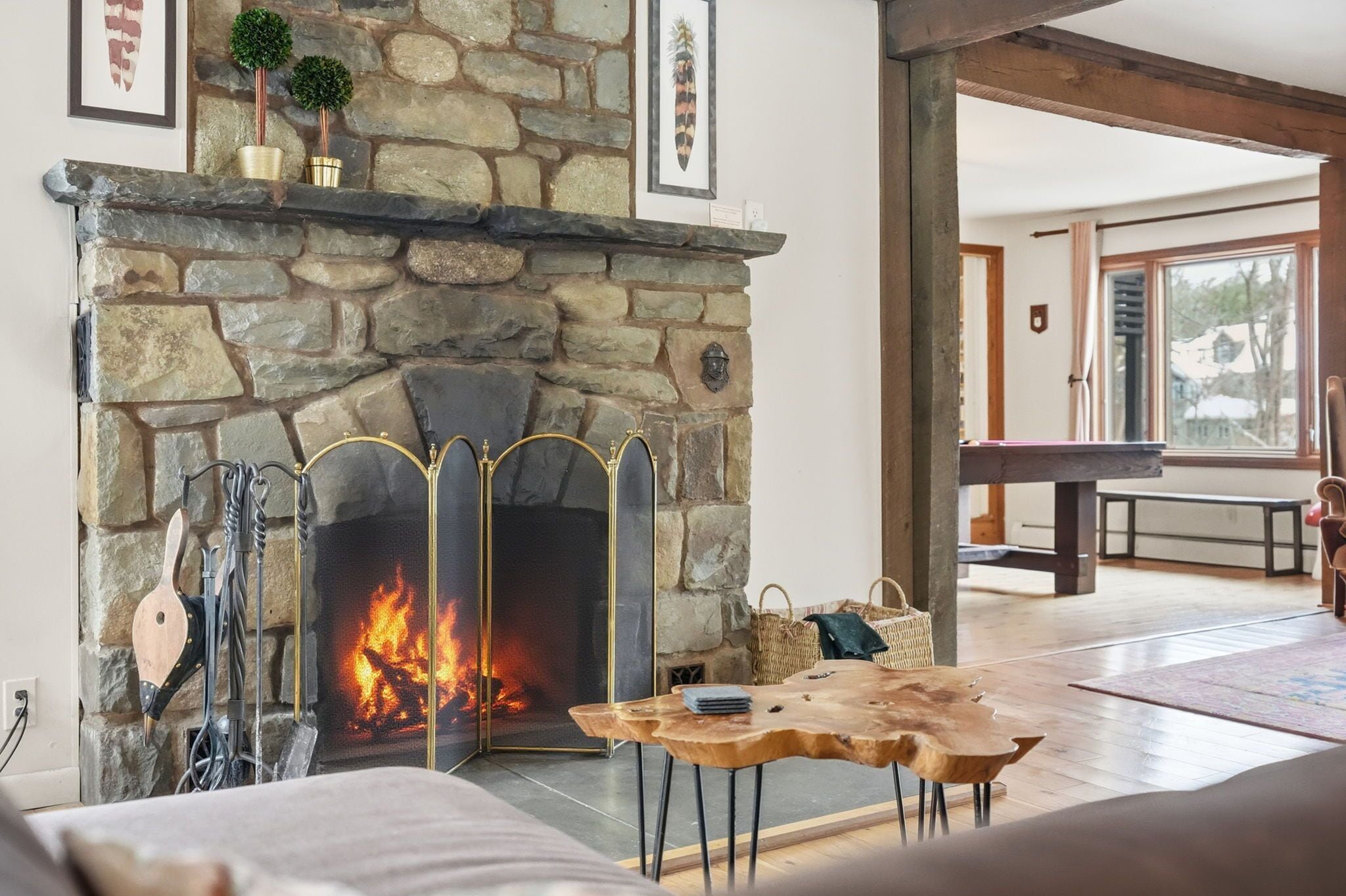 A stone fireplace with a fire, a wooden table, and a bench in a living room with a view of the outdoors.