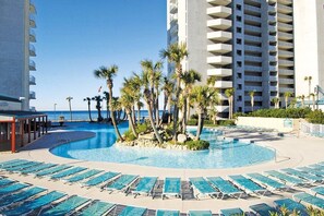 A resort pool area with multiple sun loungers and palm trees, surrounded by high-rise buildings.