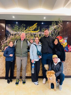 A group of people posing in front of a reception desk with a decorative floral arrangement and a sign.