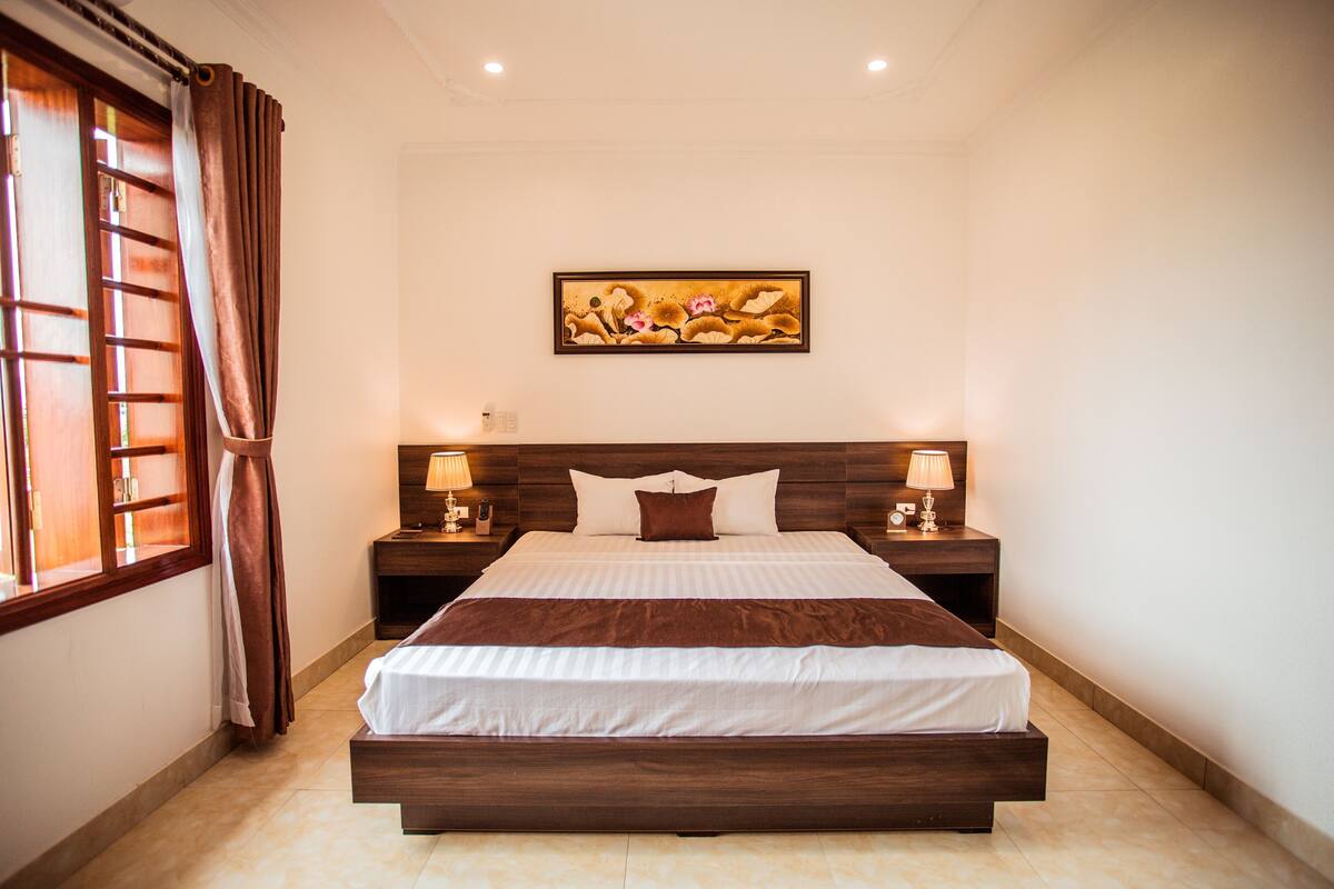 A bedroom with a large bed, wooden headboard, two bedside tables with lamps, a window with curtains, and a painting on the wall.