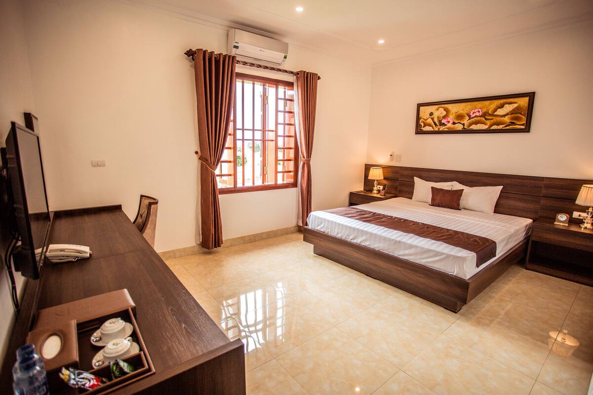 A hotel room with a bed, bedside tables, a desk with a telephone, a mirror, and an air conditioning unit.