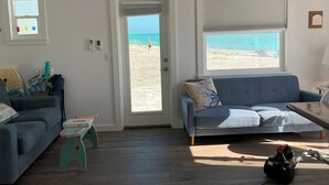 Ein Wohnzimmer mit Blick auf den Strand, eine blaue Couch, ein kleiner Tisch und ein Staubsauger auf dem Boden.