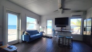 Ein Zimmer mit einem blauen Sofa, einem an der Wand montierten Fernseher, einem Deckenventilator und großen Fenstern mit Blick auf den Strand.