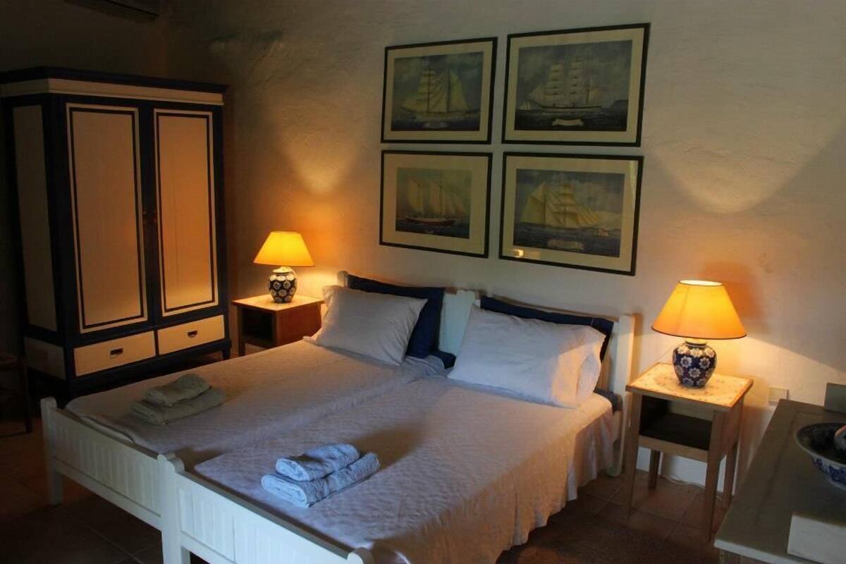 Ein ordentlich bezogenes Bett mit weißen Laken, zwei Nachttische mit Lampen, ein Kleiderschrank und vier gerahmte Bilder an der Wand.