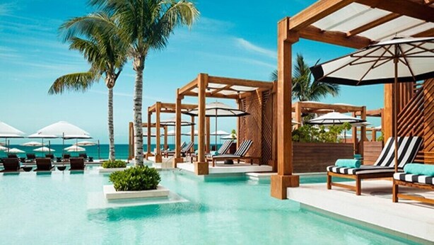 A pool area with lounge chairs and umbrellas, surrounded by wooden pergolas and palm trees.