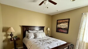A bedroom with a wooden bed, a ceiling fan, a bedside table with a lamp, and a painting on the wall.