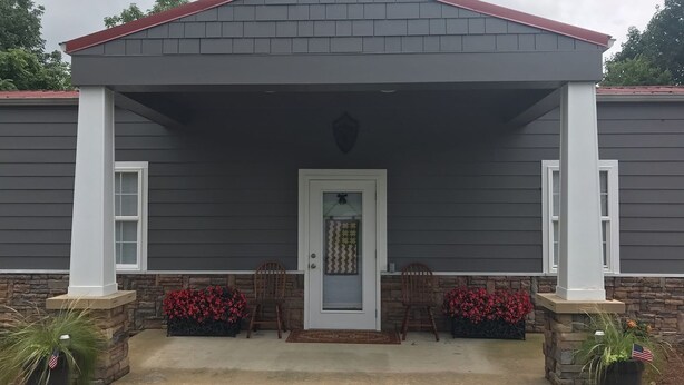 A two-story house with a grey exterior and a red roof. It features a front porch with two chairs and a small table, and a door with a decorative glass panel.