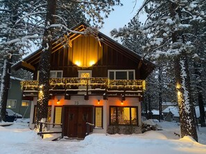 Een tweelaags lodge met balkon, besneeuwde bomen en een sfeervol verlichte ingang.