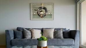 A living room with a grey sofa, patterned cushions, a glass coffee table, and a decorative wreath on the wall. - Oceanfront Brand New Unit at La Jolla Excellence
The Pelican  (Playa Encantada)