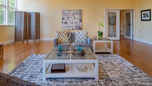 A spacious living room with a large window, a sofa with patterned cushions, a coffee table with decorative items, and a painting on the wall. - Sleeps 10 + Spa + Foosball | 3 Acres of Rural Privacy | Union Road Schoolhouse (Paso Robles)