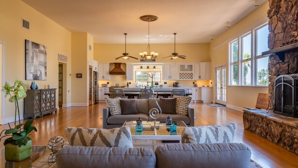 A spacious living room with a sofa, coffee table, and decorative items. A kitchen is visible in the background with white cabinets and a stone fireplace. - Sleeps 10 + Spa + Foosball | 3 Acres of Rural Privacy | Union Road Schoolhouse (Paso Robles)