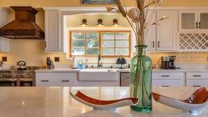 A kitchen with white cabinets, a stainless steel stove, and a window with a view of trees. - Sleeps 10 + Spa + Foosball | 3 Acres of Rural Privacy | Union Road Schoolhouse (Paso Robles)
