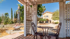 A covered patio with two metal chairs and a table, overlooking a landscaped garden with a white wheelbarrow of flowers. - Sleeps 10 + Spa + Foosball | 3 Acres of Rural Privacy | Union Road Schoolhouse (Paso Robles)