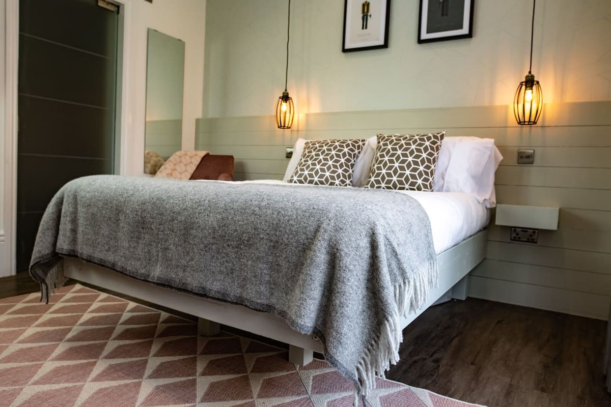 Un dormitorio con cama, almohadones estampados, una manta gris y dos lámparas fijadas a la pared.